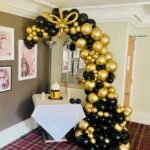 Demi Arch with decorative flower balloon . Mirror Gold and Standard Black organic style with a thicker base tampering towards the end over a cake table .